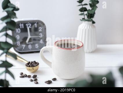 Breakfast relax time concept. coffee mug on a cozy kitchen table with alarm clock and green plants in vase. scandinavian minimal style. coffee beans in a spoon. time for yourself to start a good day. Stock Photo