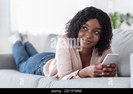 Pensive african woman using smartphone while lying on comfy sofa at home Stock Photo