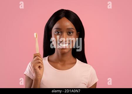 Portrait of beautiful African American woman with radiant smile holding wooden toothbrush with paste on pink background Stock Photo