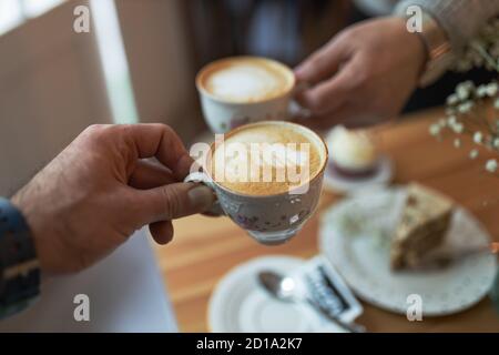 Closeup shot of hands holding cups of cappuccino with foam drawing over the table Stock Photo
