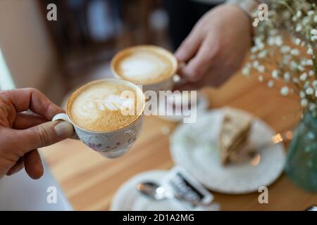 Closeup shot of hands holding cups of cappuccino with foam drawing over the table Stock Photo