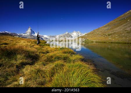 Hikers hiking the Five Lakes trail with the Matterhorn in the background, one showing an angler fishing in the Stellisee. Stock Photo