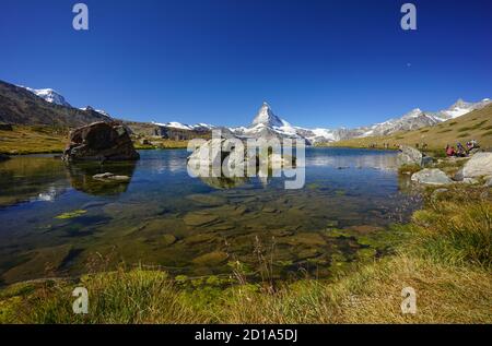 Hikers hiking the Five Lakes trail with the Matterhorn in the background, one showing an angler fishing in the Stellisee. Stock Photo