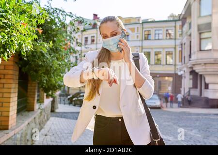 Young business woman in medical protective mask talking on smartphone, looking at wristwatch Stock Photo