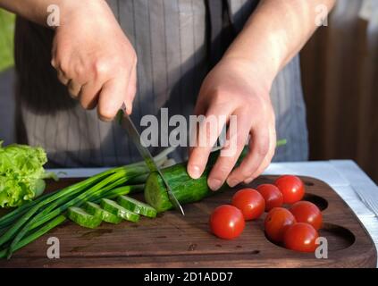 woman in grey apron cutting vegetables for salad. cropped view of woman hands holding knife and making salad. cucumber, cherry tomatoes and green onion on the table. Stock Photo