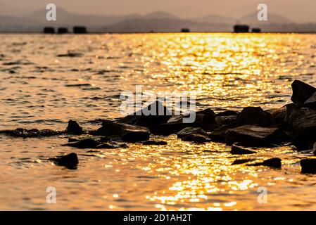 Beautiful nature landscape of bright golden sunlight reflects the yellow glow on the ripples pile of rock in the water of a tropical lake mountain bac Stock Photo