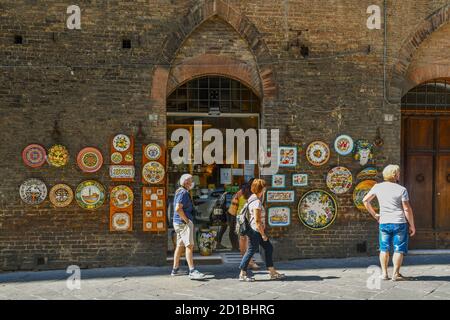 Exterior of a handmade ceramics shop with decorated plates hanging on the old brick wall and tourists in the historic centre of Siena, Tuscany, Italy Stock Photo