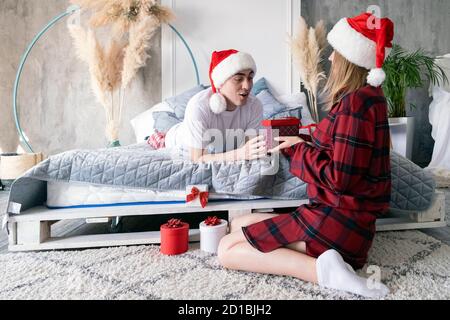 Christmas congratulations. Excited young woman with gift present box wearing santa hat. Home holiday, happy couple Stock Photo
