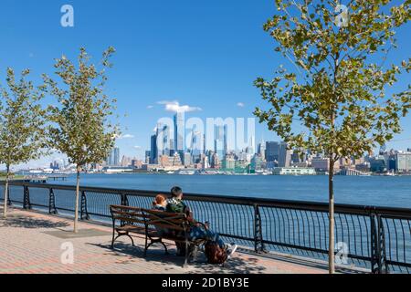 Couple sitting on bench enjoying view of New York City/Manhattan skyline from across the Hudson River at Hoboken, New Jersey's waterfront. Stock Photo