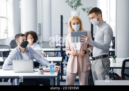 Teamwork in corporate company and social distance during coronavirus epidemic. Male manager in protective mask shows tablet to workers in interior Stock Photo
