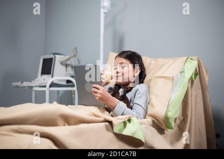 Teenage Female Patient Relaxing In Hospital Bed With Digital Tablet. Stock Photo