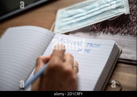 Working from home during Covid 19 global pandemic conceptual image - female hand writing in notepad with medical protective mask next to it. Stock Photo
