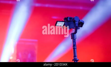 Digital mirrorless camera on handheld gimbal stabilizer taking photo or recording video of live music concert with red illumination. Photography Stock Photo