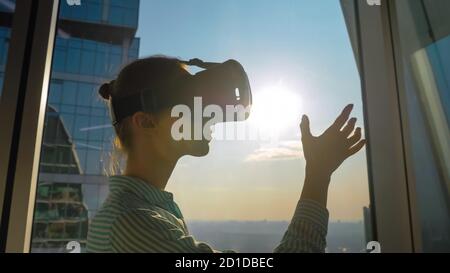 VR, sightseeing, discover, technology concept. Woman using virtual reality headset and moving hand against view on cityscape from panoramic skyscraper Stock Photo