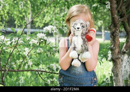 cute little girl hiding behind toy cat. caucasian blonde girl hides her face. playing outdoors in a beautiful place near blooming white tree. life after quarantine.