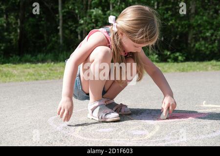 happy little girl drawing with chalk on the pavement. blonde caucasian girl draws in the park, life after quarantine. active games outdoors. Stock Photo