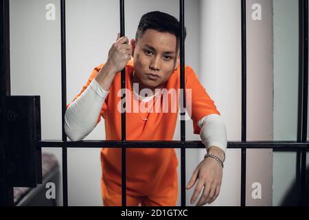 A convicted asian man in an orange robe, with a chain and bracelet and a tattoo on his face stands in a prison cell next to the bars Stock Photo