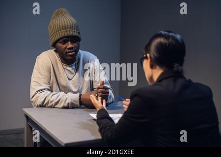 A beautiful girl investigator in an elegant suit asks a black criminal to cooperate with the investigation and sign a confession Stock Photo