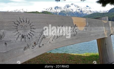 St. Moritz, Graubünden, Switzerland - 30th September 2020 : St. Moritz sign engraved on a wooden bench with snowy mountains and lake in the background Stock Photo
