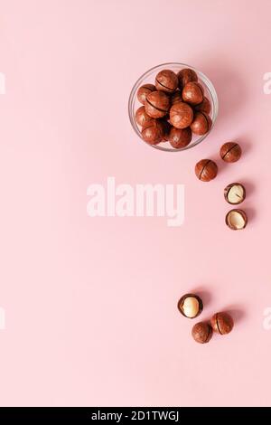 Flat lay composition with raw macadamia nuts in a transparent glass bowl and scattered on a pink solid background. Minimal composition. Stock Photo