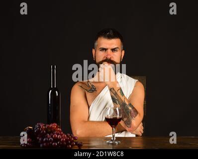 Winemaking and degustation concept. Man with beard near glass of alcohol on brown background. Sommelier tasting drink. God Bacchus with thoughtful face in white cloth sits by wine bottle and grapes Stock Photo