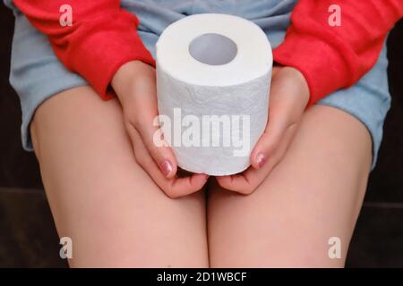 A woman sits on the toilet bowl and holds a roll of soft paper in front of her, close-up. Concept of abdominal disease and problems with diarrhea. Stock Photo