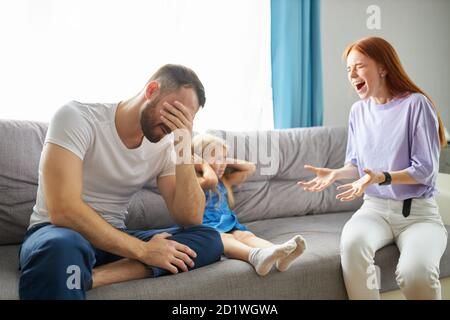 sad, desperate little girl during parents quarrel, she clog the ears sitting on sofa at home, angry parents fighting. worried upset small daughter hurt by fathers and mothers break up or divorce Stock Photo