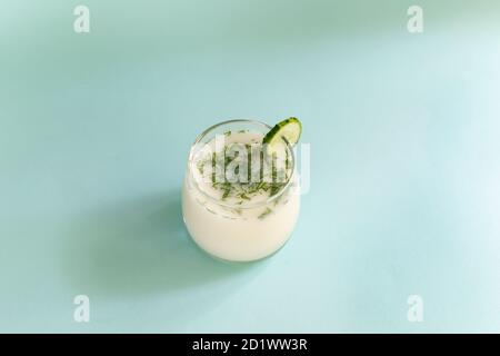 Ayran in a glass glass with dill and a slice of cucumber on a blue background. Fermented product concept. Copy space. Stock Photo