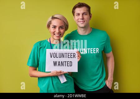 two caucasian man and woman waiting for new people in their charity organization, they hold paper with description volunteers wanted Stock Photo