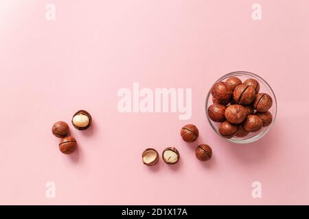 Raw macadamia nuts in a transparent glass bowl and scattered on a pink solid background. Minimal composition. Stock Photo