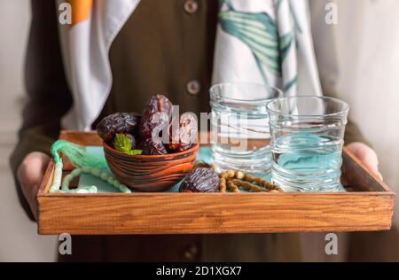 A female hand holds a tray with dates, and water and a rosary - hthe things used to break the fast at sunset during the Muslim holy month of Ramadan. Stock Photo