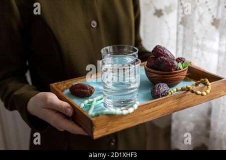 A female hand holds a tray with dates, and water and a rosary - hthe things used to break the fast at sunset during the Muslim holy month of Ramadan. Stock Photo