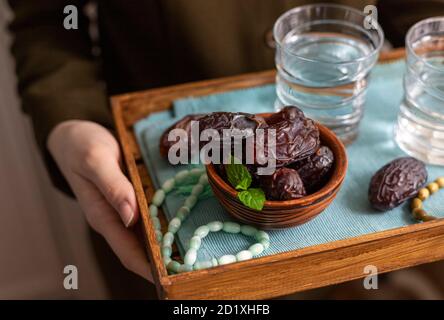 A female hand holds a tray with dates, and water and a rosary - hthe things used to break the fast at sunset during the Muslim holy month of Ramadan. Stock Photo