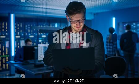 Portrait of a Smart Focused Young Man Wearing Glasses Holds Laptop. In the Background Technical Department Office with Specialists Working and Stock Photo