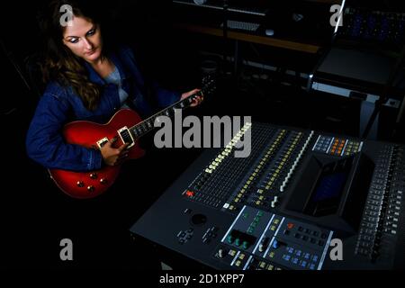 Female guitarist sitting at the mixing console in the recording studio. Woman with a red electric guitar records a song at the music studio. Stock Photo