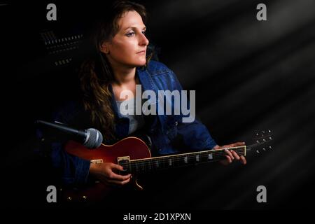 Woman with a red electric guitar in a denim jacket records a song at a music Studio. Professional guitarist behind a microphone on a black background. Stock Photo