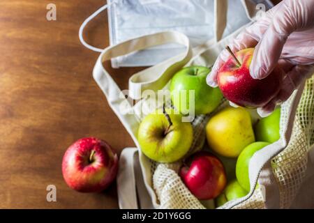 Hand in glove holds apple. Eco mesh bag with food supplies on a wooden background. Food delivery in eco-friendly packaging, zero waste. Stock Photo