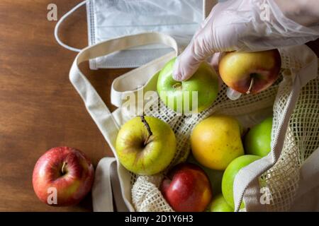 Hand in glove holds apple. Eco mesh bag with food supplies on a wooden background. Food delivery in eco-friendly packaging, zero waste. Stock Photo