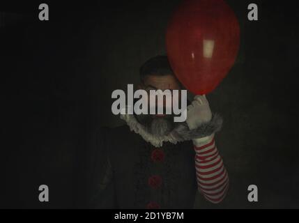 Evil and scary man holding red balloon in hand on black background .Halloween vintage costume. Painted Halloween holiday makeup. Stock Photo
