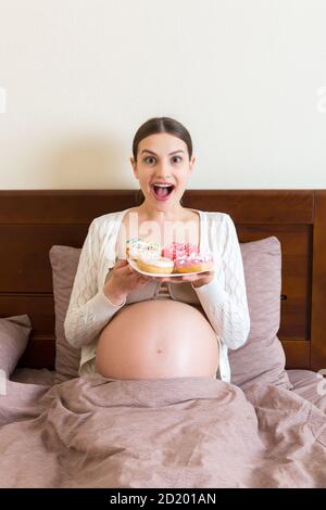 Pregnant woman is eating many donuts relaxing in bed. Unhealthy dieting during pregnancy concept. Stock Photo