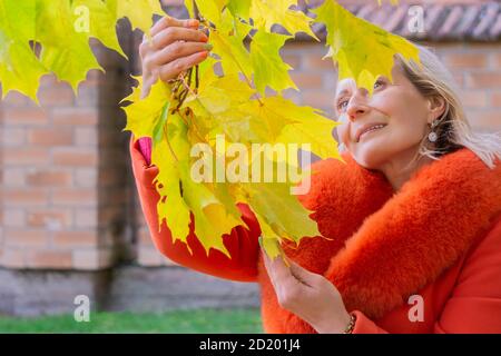 Portrait of middle-aged woman in orange coat holding autumn maple leaves branch in red brick wall background. Portrait of romantic female Stock Photo
