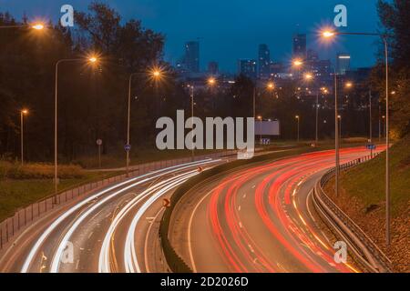 Cars light trails on a curved highway to the city at night. Night traffic trails and cityscape. Light city road with traffic headlight motion. Stock Photo