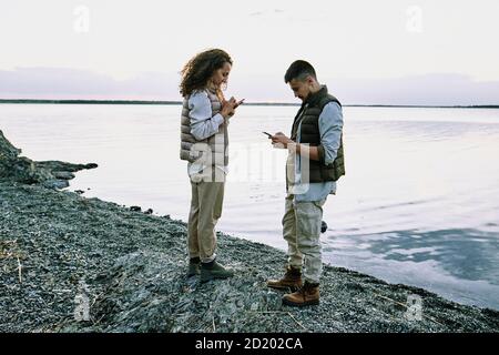Concentrated young couple standing on pebble beach and using gadgets while checking connection Stock Photo