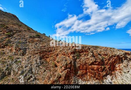 Beautiful wild red cliffs, clear blue sky, clouds. Diagonal view. Typical Greek landscape. Akrotiri peninsula, Chania region, Crete island, Greece Stock Photo