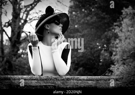Young Caucasian woman in 1950s Audrey Hepburn styling, bodycon black and white dress, wide brimmed black hat, black sunglasses and a cigarette holder. Stock Photo