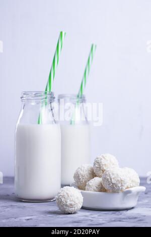 Milk in glass bottles with green straws with white coconut round sweets on gray table on white background Stock Photo