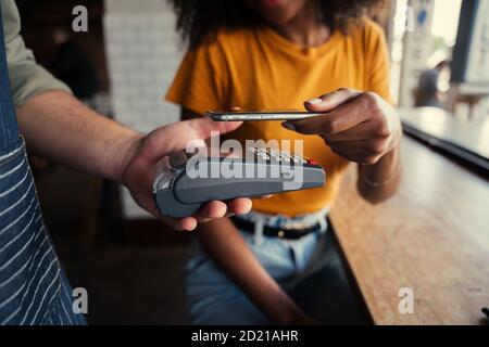 African American female scanning bar code of card machine with smartphone paying for coffee in funky cafe. Stock Photo