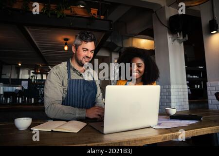 Male and mixed race female coffee shop owners smiling at laptop in coffee shop. Stock Photo