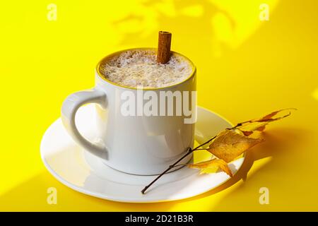 Autumn coffee or cappuccino composition. White coffee cup with milk foam, cinnamon stick and autumn leaves on yellow background. Fall hot drinks, cafe Stock Photo