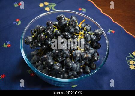 Black grapes in a glass bowl on the background of a wooden table covered with a tablecloth close-up. Stock Photo